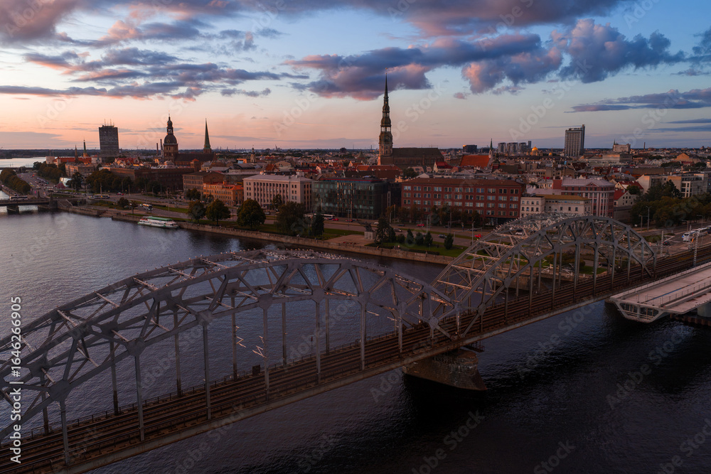 Fototapeta premium Riga, Latvia at sunset with the Daugava River, Railway Bridge, and Old Town landmarks like St. Peter's Church and Riga Cathedral in the background.