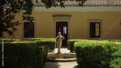 Stone statue and fountain in a manicured garden before a yellow Mediterranean villa