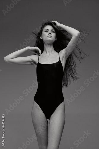 Black and white portrait of a slender woman posing in a swimsuit with flowing hair and confident expression against a plain background.