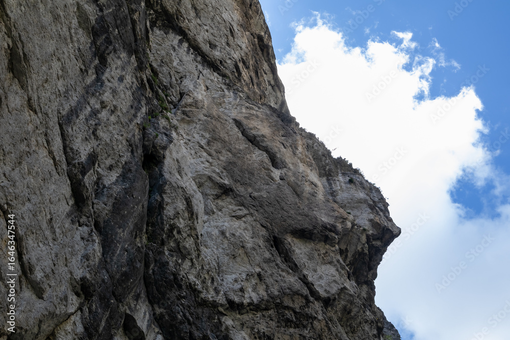 A dramatic low-angle view of a towering limestone cliff face in Romania's Bicaz Canyon (Cheile Bicazului), with intricate rock textures set against a bright blue sky with white clouds. A dramatic low-angle view of a towering limestone cliff face in Romania's Bicaz Canyon (Cheile Bicazului), with intricate rock textures set against a bright blue sky with white clouds.
