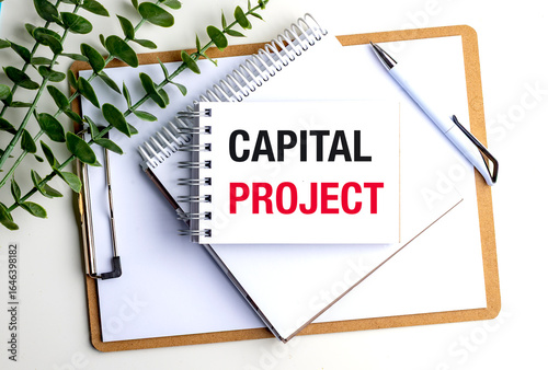 Notepad with capital project text on clipboard with pen and green plant