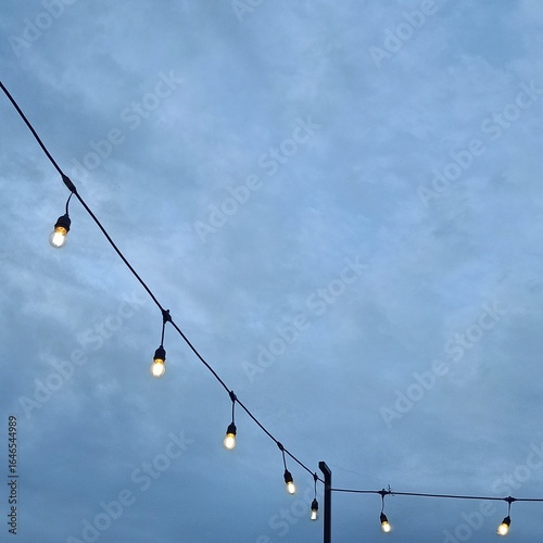 A string of hanging light bulbs glowing under a cloudy evening sky.