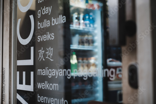 Multilingual welcome sign decal  on the transparent glass door of a tourist spot kiosk selling cold beverages with a refrigerator seen on the background