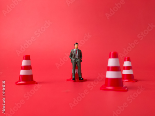 The image features a miniature figurine of a person standing on a traffic cone, with three other traffic cones on a red background