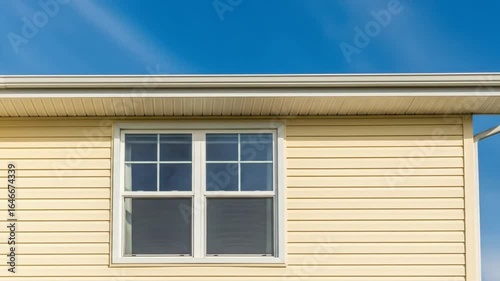 Exterior of a residential house with yellow vinyl siding, a white window, and a roof gutter against a clear blue sky.