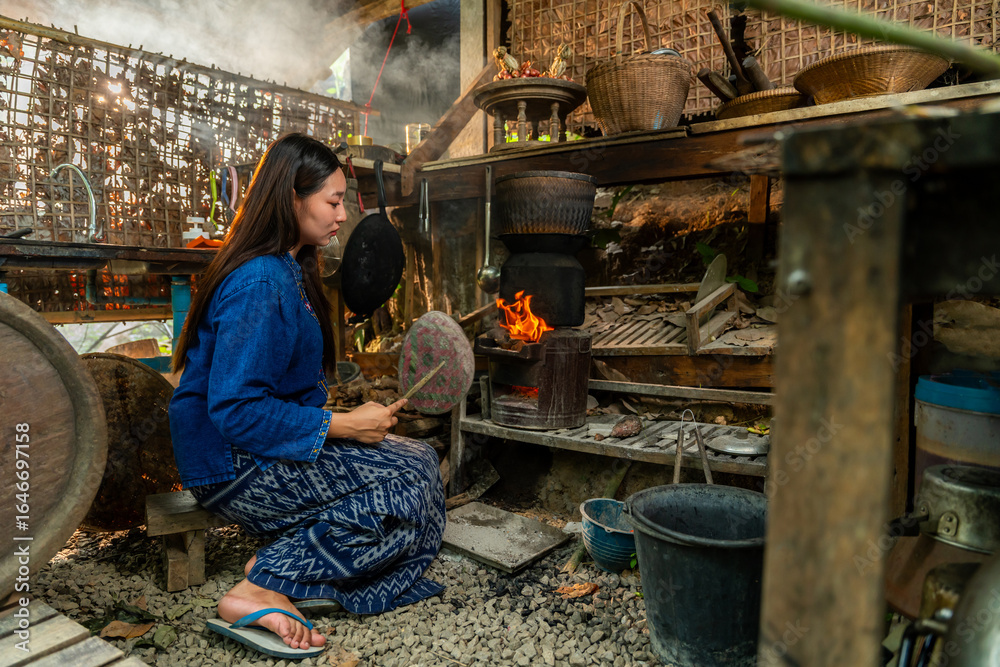 Obraz premium A Thai woman in traditional attire fans a charcoal stove in a rustic kitchen, capturing the warmth and charm of local culture.