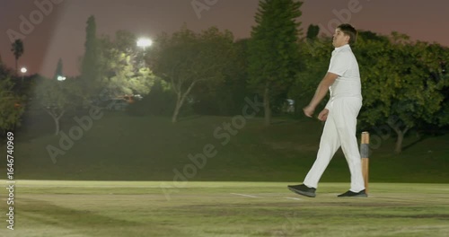 Delivering ball male bowler watching batsman leave crease resetting run-up on grass pitch in lights