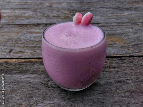 Strawberry milkshake with smooth foam in rounded glass, topped with pink-white gummy candies, served on rustic wooden table. 
