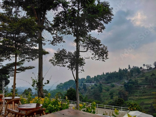 Scenic hillside view with outdoor wooden seating, lush green landscape, and cloudy sky.