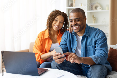 Foto Smiling African American couple using smartphone together at home, sitting at table with laptop, looking at screen and enjoying moment