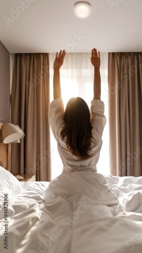 Woman stretches arms wide in bed, back view, bathed in morning light