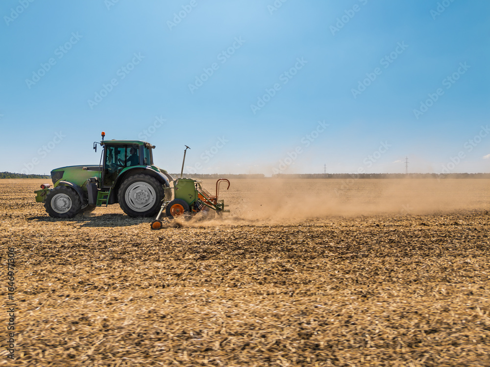 Fototapeta premium Green tractor plowing dry field on a sunny day, raising dust