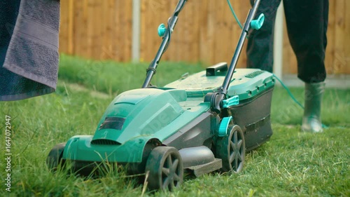 Wallpaper Mural Operator pushing electric lawn mower over tall grass in rubber boots. Individual cutting backyard lawn using corded mower while wearing green wellingtons. Person trimming thick grass with powered Torontodigital.ca