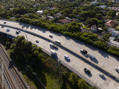 Aerial view of cars and trucks streaming along a sun-drenched highway, a concrete ribbon cutting through lush greenery and shadowed neighborhoods, Miami, Florida, United States.