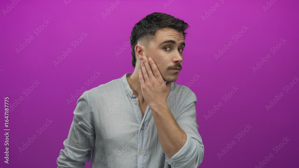 Fototapeta premium Young hispanic man with short hairstyle touching his face and hair while standing in front of a vibrant isolated pink wall.