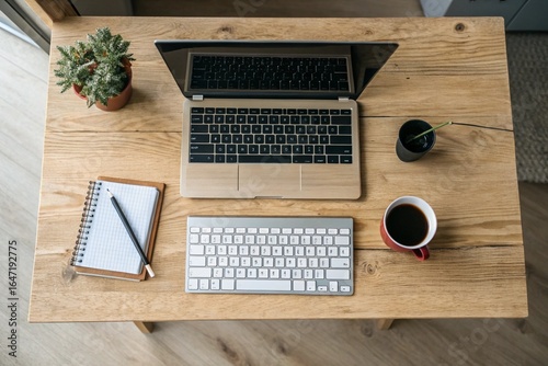 Home Office Desk Setup: Overhead view of a laptop, wireless keyboard, coffee mug, and a small plant on a light wooden desk