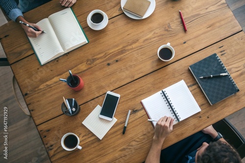 Office Meeting Table: Overhead shot of a few notebooks, coffee cups, and pens spread out on a large wooden table