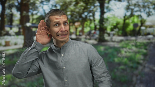 Caucasian man listening with hand to ear and expressing emotions in an outdoor park setting.