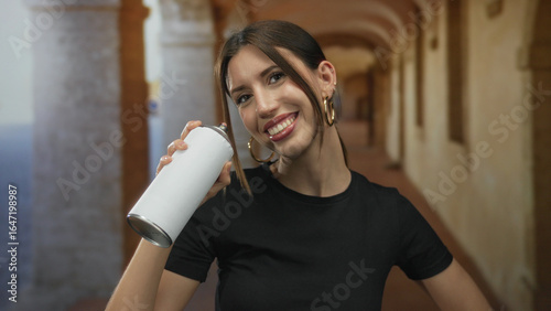 Photography Woman holds white spray can with hand while smiling in a historic building corridor; urban creativity