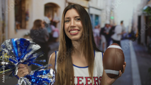 Foto Cheerleader smiling confidently holds silver pom pom and brown football on urban street; enthusiasm
