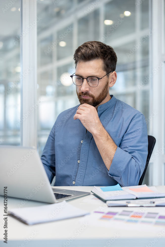 Fototapeta premium A professional individual works on a laptop in a modern, bright office environment. They appear thoughtful, concentrated, and engaged with their tasks, surrounded by organized office essentials.