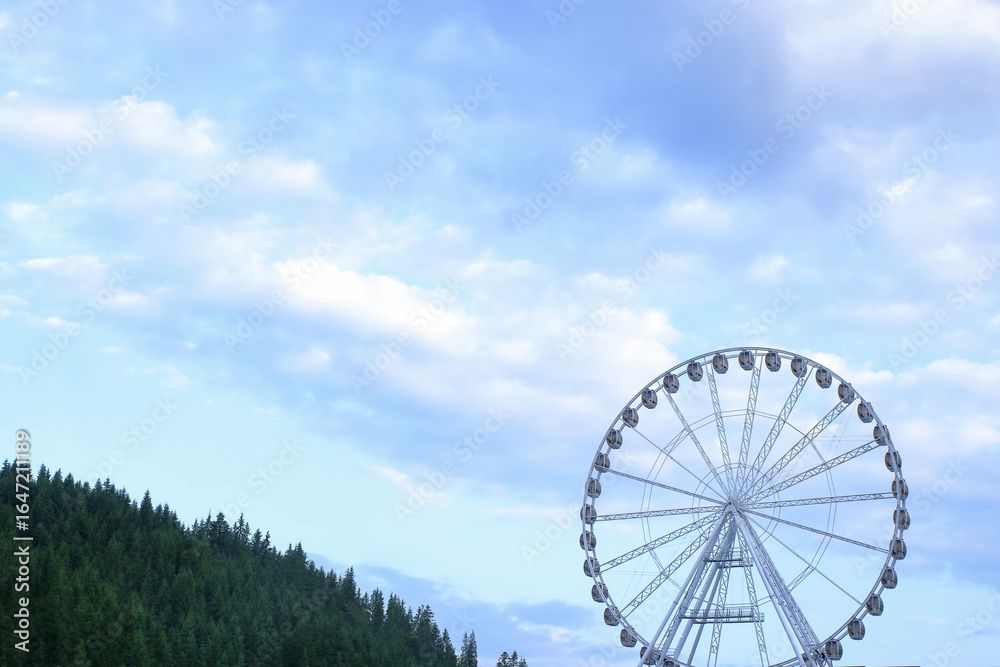 Fototapeta premium White Ferris wheel rising above lush green forest with mountain backdrop, scenic summer view, travel and leisure destination, eco tourism. Ferris wheel stands tall against a deep blue sky