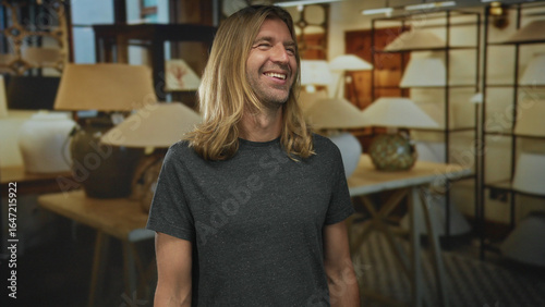 Blond man smiling beside lamp display on wooden table inside a building with shelves in view; happiness.
