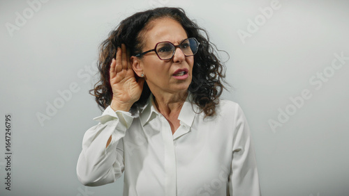 Woman with curly hair and glasses, hand to ear in surprise against a white background, showcasing attentive listening or surprise in a neutral setting.