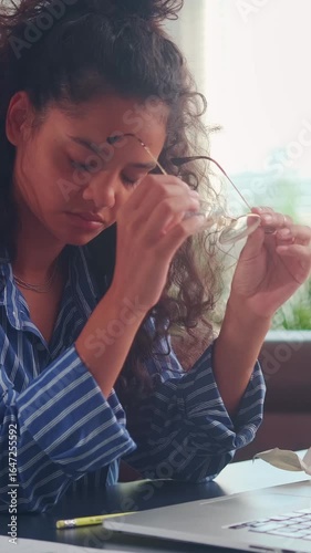 Young Indian woman sits at her desk, surrounded by crumpled papers, struggling with her work. Frustrated and exhausted, she removes her glasses and rubs her temples, fighting through the pressure.