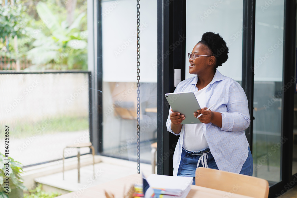 Obraz premium Businesswoman is holding a tablet and stylus while looking out the window during a work break, taking a moment to relax and enjoy the view of the garden outside