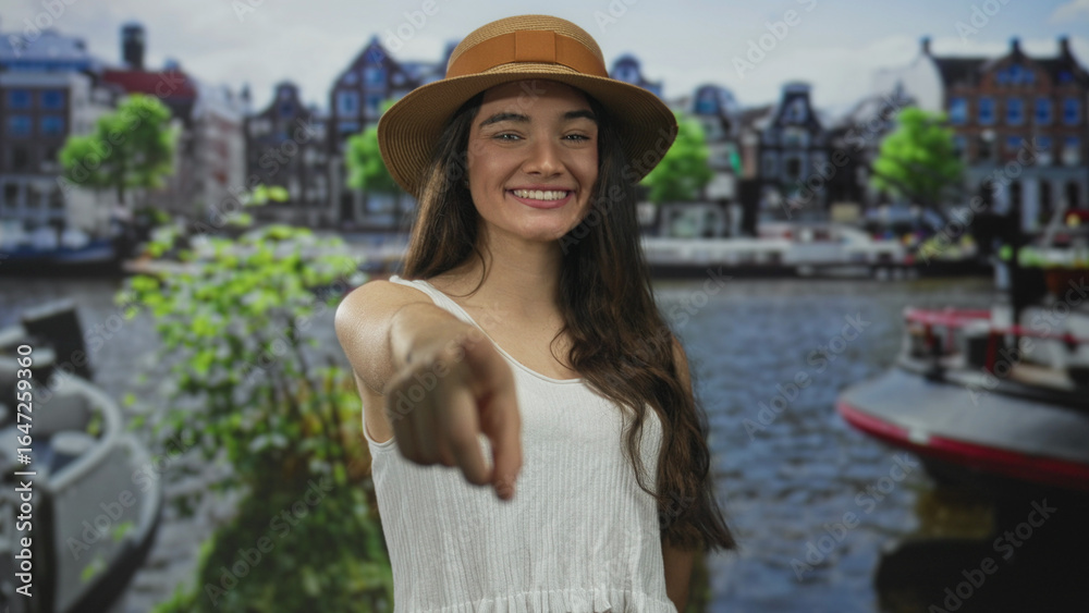 Fototapeta premium Hispanic teenager wearing summer hat smiles and points finger forward in street; joyful connection invitation.