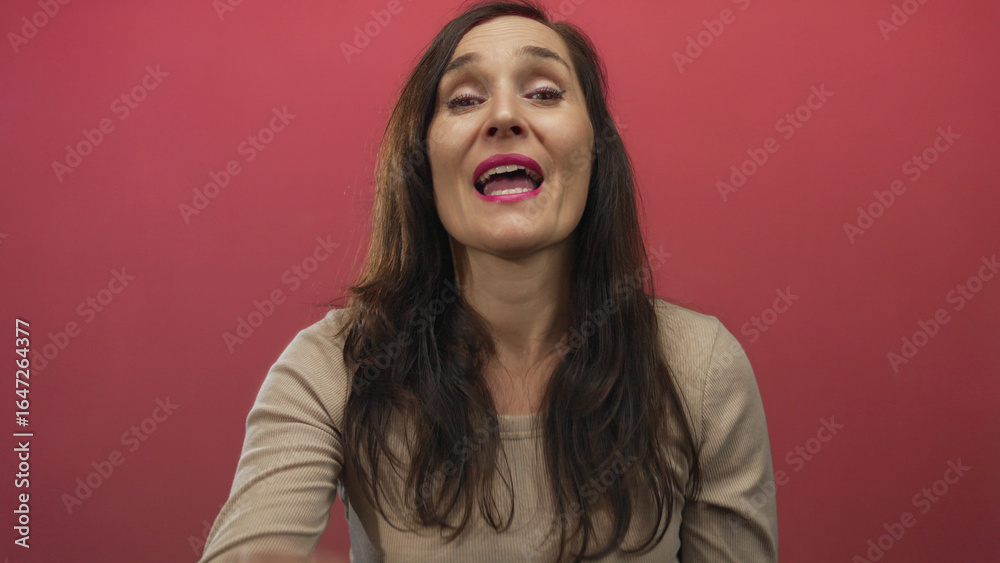 Fototapeta premium Hispanic young woman cups hand to mouth shouting against red wall; urgent alert announcement expression.