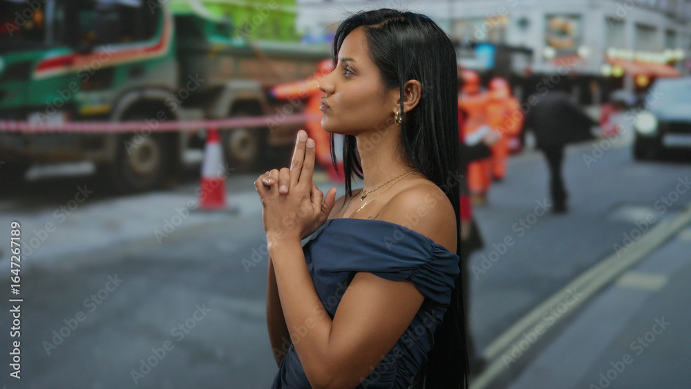Fototapeta premium Young woman makes hand gesture resembling a gun at a city construction site with blurred workers and vehicles in the background.