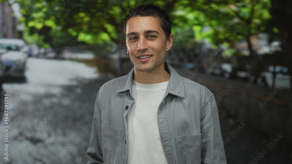 Young man standing outdoors on a sunny urban street with trees in the background, wearing casual clothing and smiling in an urban town environment. Young man standing outdoors on a sunny urban street with trees in the background, wearing casual clothing and smiling in an urban town environment.