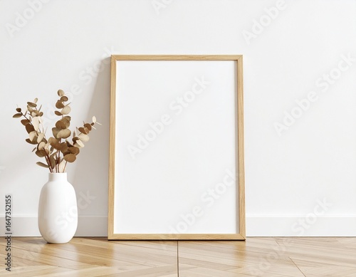 Minimalist interior scene with a white vase of dried eucalyptus and a wooden picture frame on a herringbone floor.