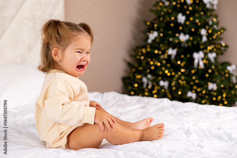 Fototapeta premium Sad and depressed toddler girl in casual bodysuit sits on a bed in front of a blurred Christmas tree background and crying. Childhood and emotions concept