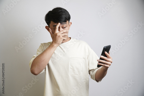 A shocked Asian man in a beige t-shirt covers his face with his hand while looking at his smartphone, reacting to something surprising or embarrassing.
