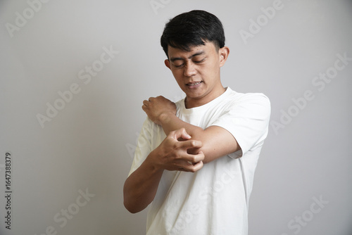A young Asian man with a pained expression, scratching his arm with his other hand, showing discomfort from an itch or a skin irritation.