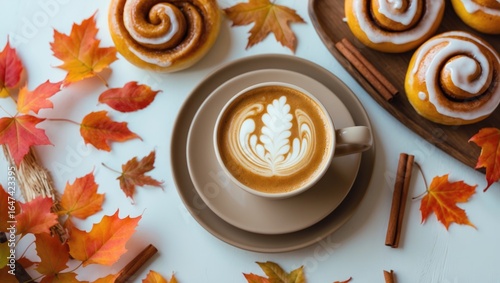 A cup of coffee with latte art, surrounded by cinnamon sticks and autumn leaves, and a plate of cinnamon rolls.
