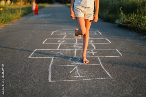 A girl plays a game of hopscotch barefoot on an asphalt road in the sunlight.