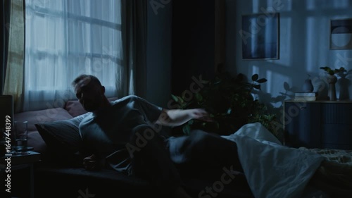 Full shot of young man suffering from PTSD twisting and jerking in bed while experiencing nightmare in dream, waking up, jumping off bed, sitting up in panic, rubbing face and drinking glass of water