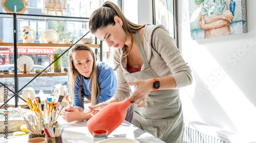 In an art studio with an environment adorned with artistic paintings in the background, two beautiful middle-aged women are joyfully painting a clay ceramic vase pink together at a table, having fun