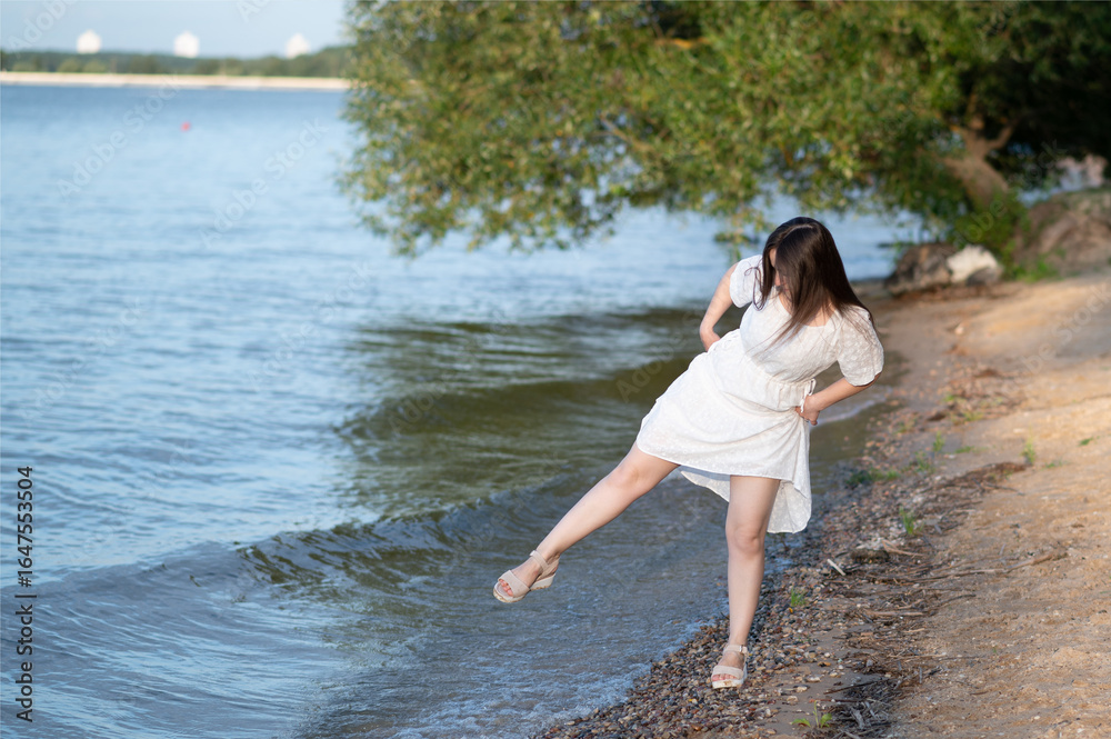 Fototapeta premium Woman in white dress walking barefoot along the shoreline, enjoying nature and water on a peaceful summer evening