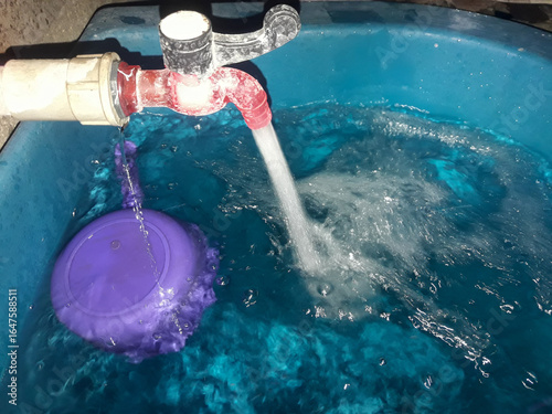 Water flows from the faucet into the bathtub. The faucet appears slightly leaky and dirty up close. Bathroom background.