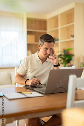 Focused middle-aged man working on laptop with a graphic tablet and pen at home office desk, thinking about project in modern bright room.
