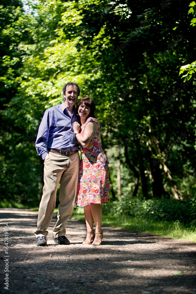 A mature, happy couple in a sun-dappled wooded park. They are smiling and looking at the camera, with the man's arm around the woman's shoulder, showcasing a loving relationship. A mature, happy couple in a sun-dappled wooded park. They are smiling and looking at the camera, with the man's arm around the woman's shoulder, showcasing a loving relationship.