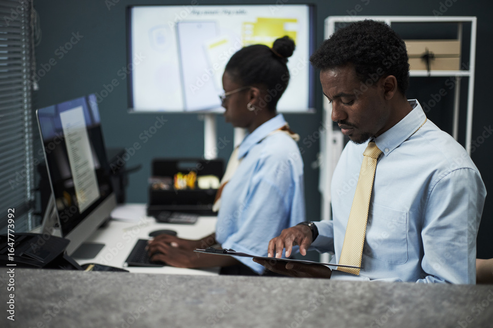 Fototapeta premium Black male bank employee reviewing documents on clipboard while Black woman working on computer in modern bank office with digital screens in background