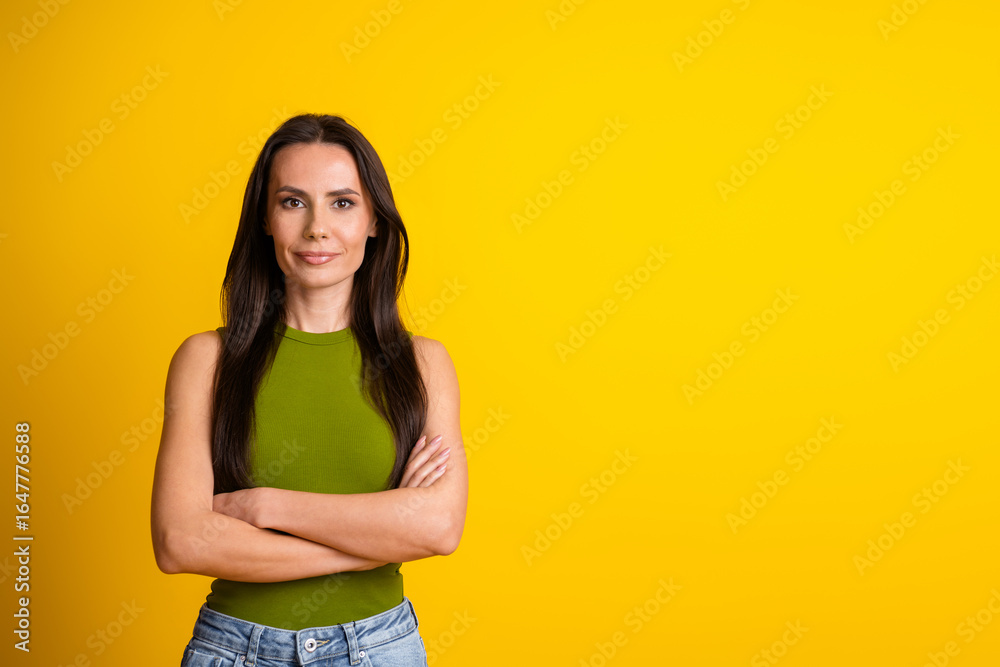 Obraz premium Confident young woman with long brunette hair standing against a yellow background in a casual green top and jeans