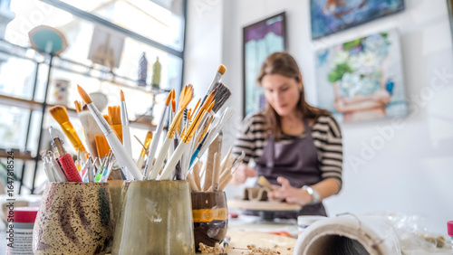 A close-up shot of paintbrushes in the foreground, with two women artists working on handmade products in the background of an art studio