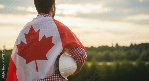 Man wearing canadian flag holding a construction helmet looking out over a scenic landscape at sunset
